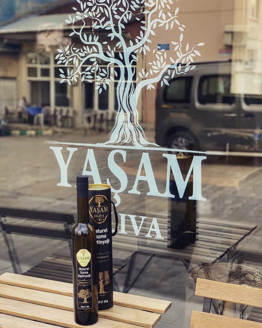 Dükkân vitrinine işlenmiş zeytin ağacı logosunun ardında duran şişe ve teneke